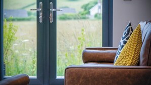 A room with a brown leather sofa, a yellow textured throw pillow, and a view of a green field through a glass door. - The Shack - 1 Bedroom - Cosheston (Pembroke)