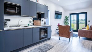 A modern kitchen with dark cabinetry, a built-in oven, and a sink. There is a dining area with a brown chair and a small table. The room has a large window with a view of the outdoors. - The Shack - 1 Bedroom - Cosheston (Pembroke)
