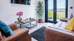 A modern living room with a sofa, coffee table, and a large window with a view of the countryside. - The Shack - 1 Bedroom - Cosheston (Pembroke)