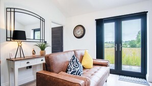 A modern living room with a leather sofa, a side table, a lamp, a clock, and a large sliding glass door leading to a view of a field. - The Shed - 1 Bedroom - Cosheston (Pembroke)