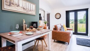A modern living room with a wooden dining table, a sofa, and a large window with a view of the countryside. - The Shed - 1 Bedroom - Cosheston (Pembroke)