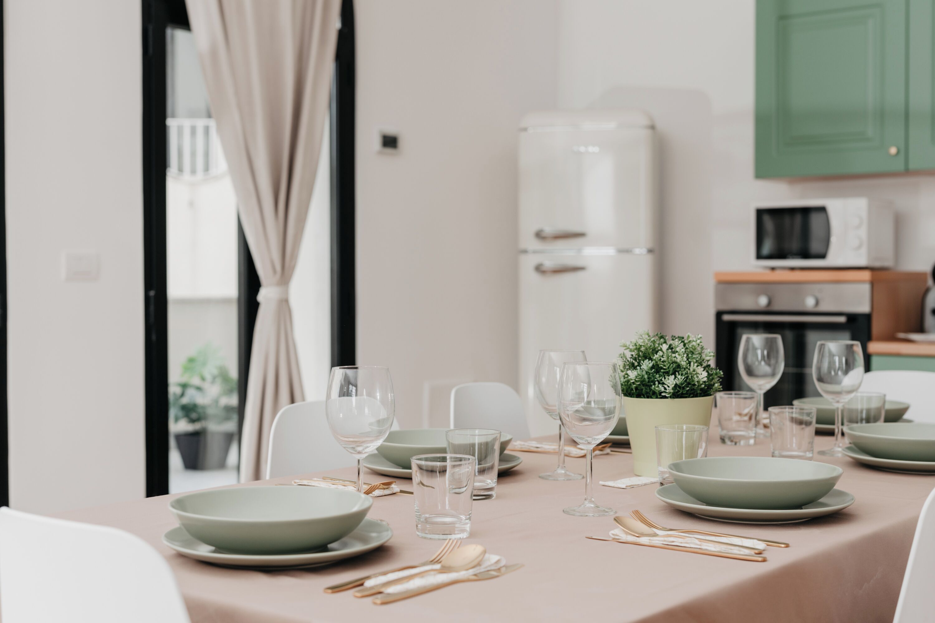 A dining table set with plates, bowls, glasses, and cutlery, with a potted plant and a microwave in the background.