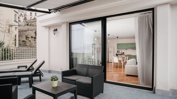 A balcony with wicker furniture, a potted plant, and a view of a kitchen and dining area through a glass door. - Vista Napoli Residence (Naples)