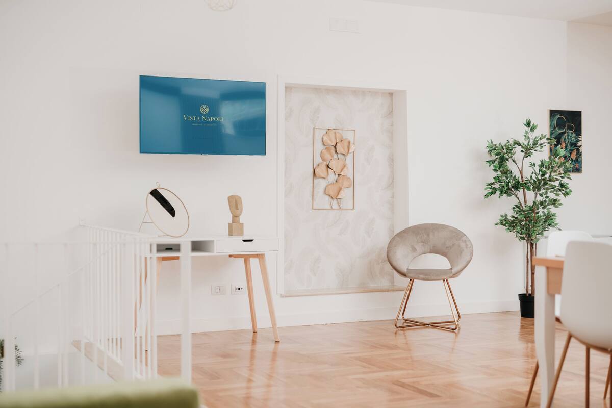 Ett modernt, minimalistiskt rum med ett vitt skrivbord, en väggmonterad TV, ett inramat konstverk och en krukväxt.