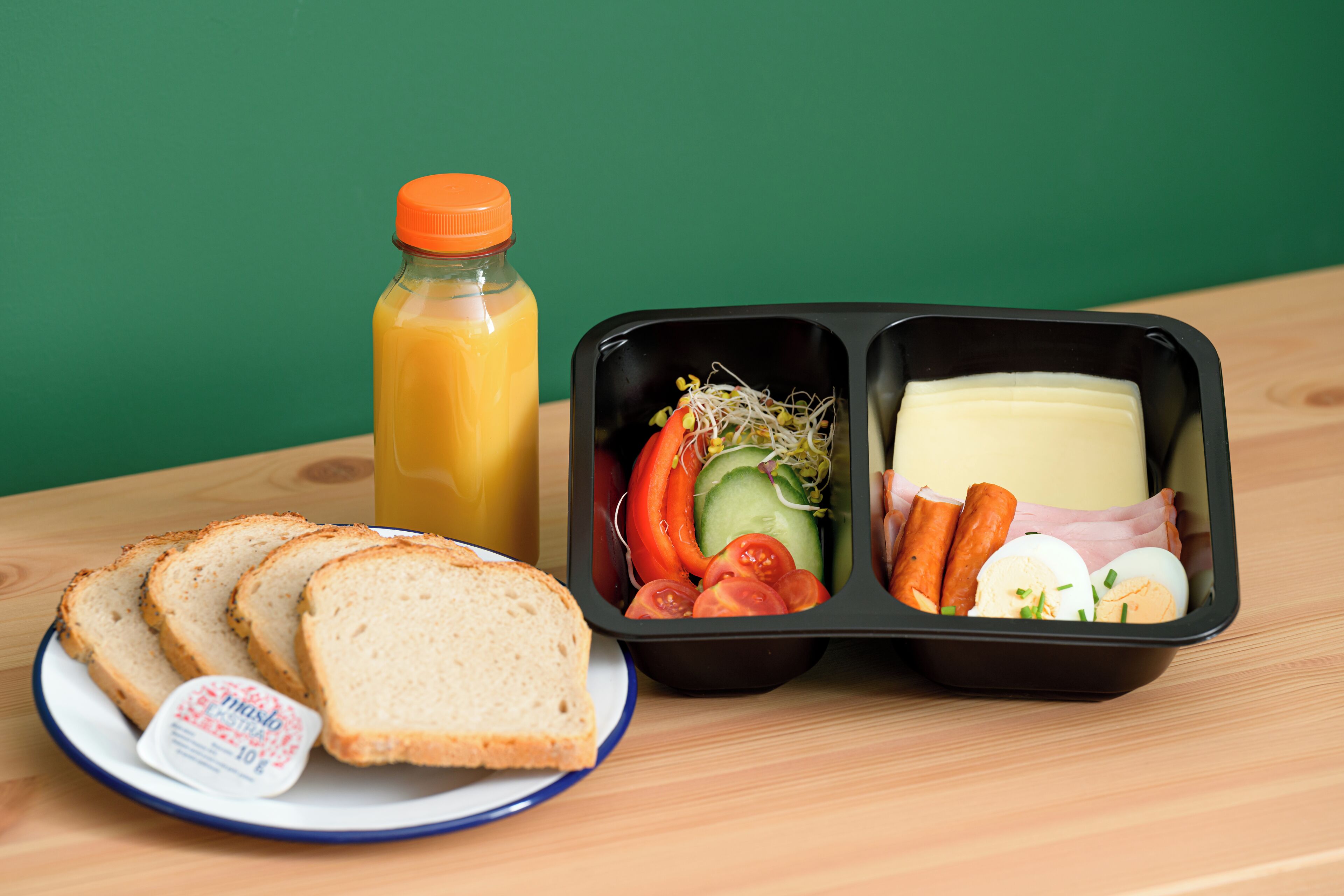 Un repas composé de pain, de tomates, de concombres, de fromage et d’œufs, le tout servi sur une assiette, accompagné d’une bouteille de jus.