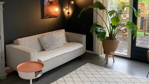 Ein modernes Wohnzimmer mit einer Couch, einem kleinen Beistelltisch, einer großen Topfpflanze und Blick ins Freie durch eine Glastür.