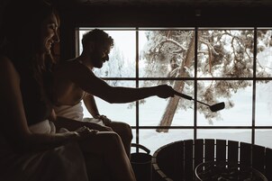 Una pareja en una habitación tenuemente iluminada con un ventanal grande, una sauna y vistas a árboles cubiertos de nieve.