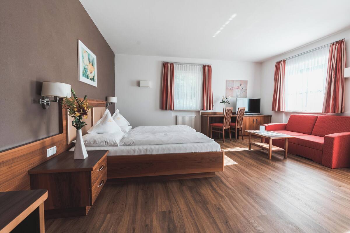 A modern hotel room with a wooden bed, a red sofa, a desk with a chair, and a large window with curtains.