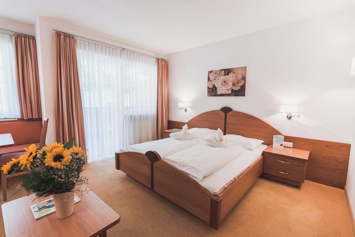 A neatly arranged hotel room with a wooden bed, a bedside table with a lamp, a small nightstand, and a wall-mounted artwork.