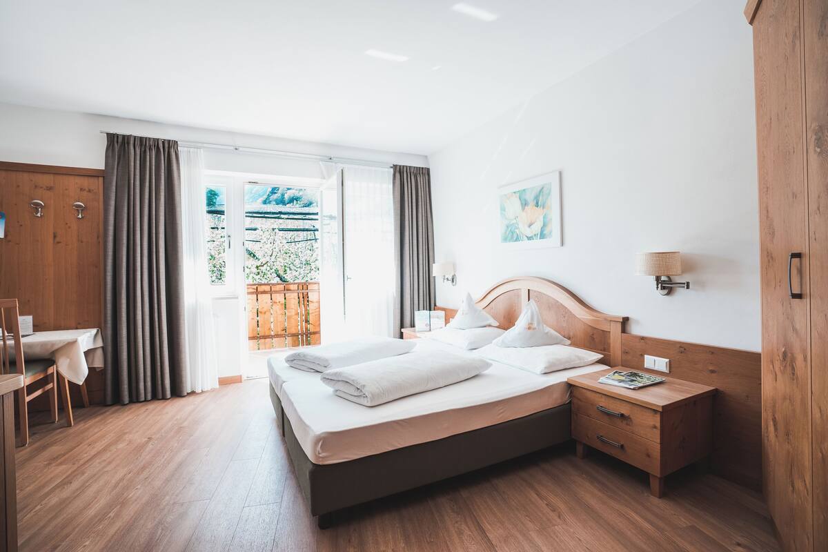 A hotel room with a wooden bed, a wooden nightstand, a desk, and a view of a garden through the window.