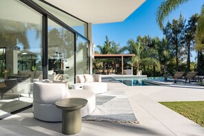 A modern outdoor living area with a pool, seating, and a glass wall. - Villa Lotus (Marbella)