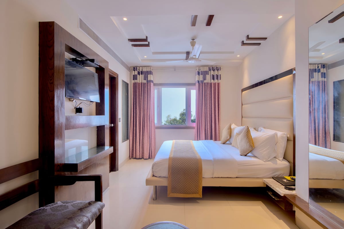 A modern hotel room with a large bed, a TV mounted on a wooden shelf, a ceiling fan, and a window with curtains.