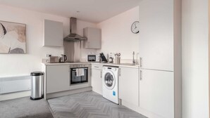 Une cuisine moderne avec des meubles blancs, une hotte de cuisinière en acier inoxydable, un four à micro-ondes, une bouilloire, un lave-linge et une poubelle.