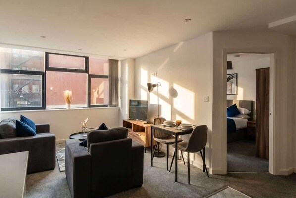 Un salon moderne avec un canapé, une table à manger et un téléviseur. Il y a une fenêtre avec des stores, une lampe et une porte qui donne sur une chambre.