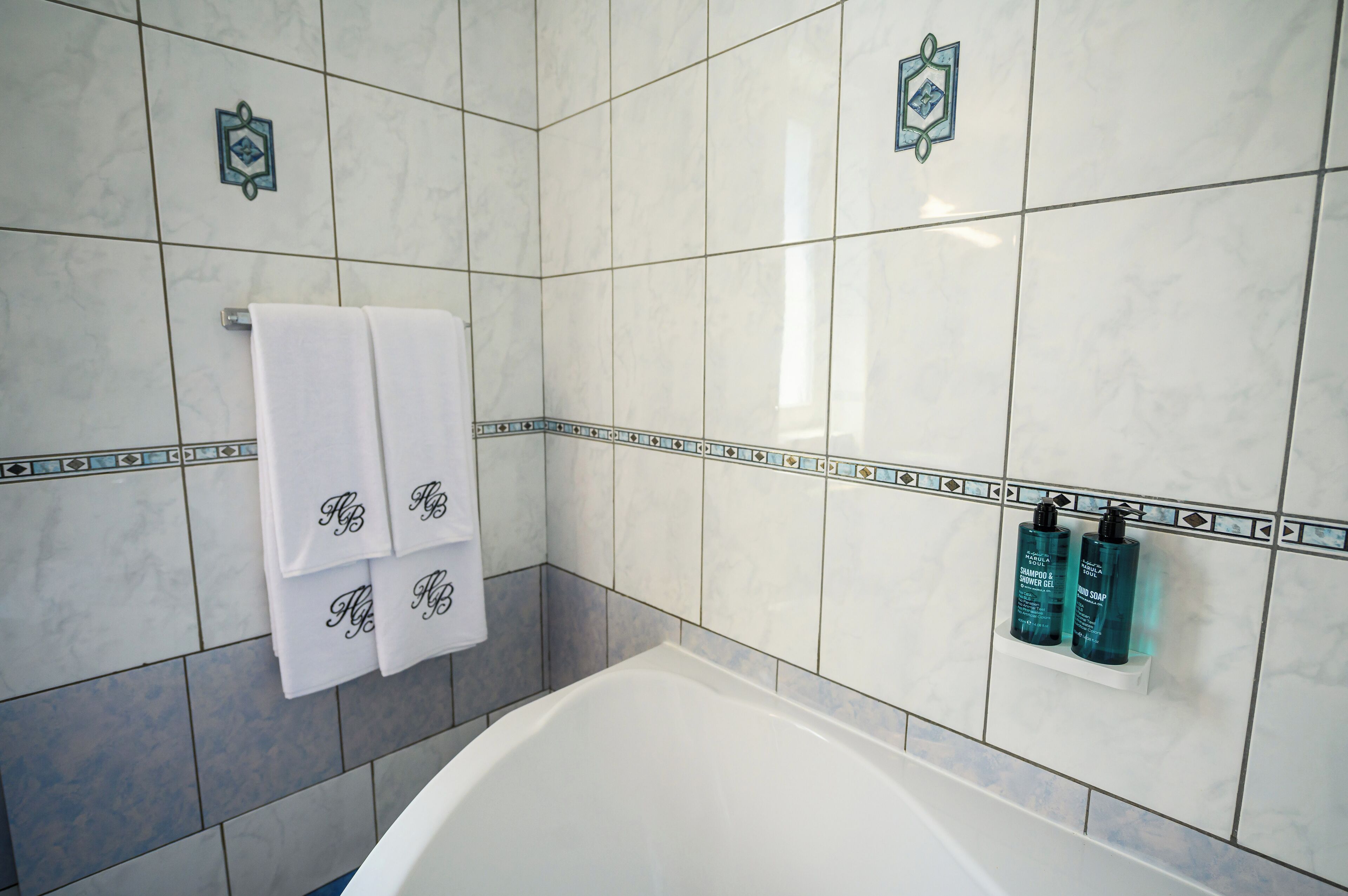 A bathroom with a white bathtub, white towels with monogrammed initials, and two bottles of shampoo and body wash on a shelf.