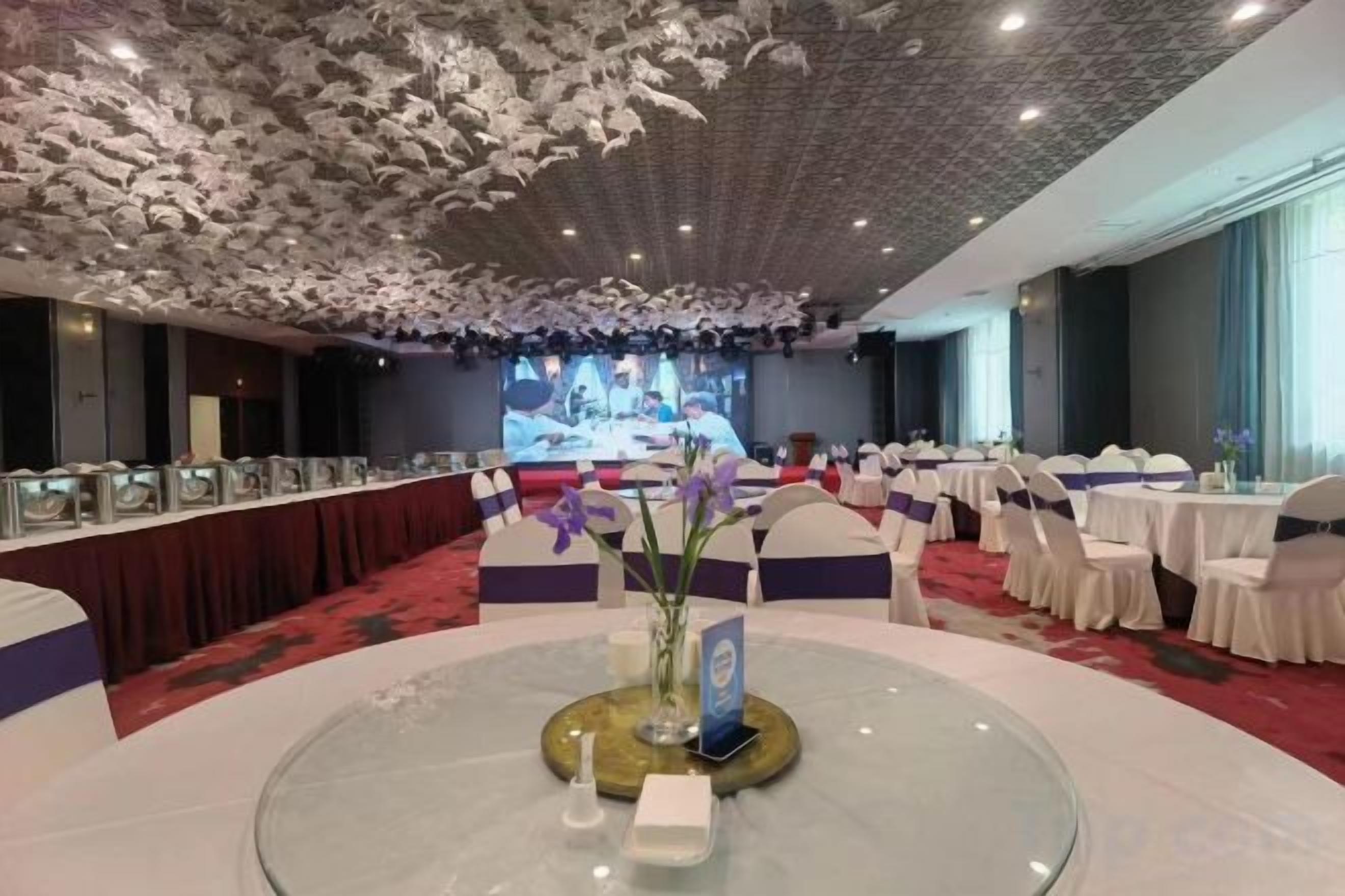 Un salón de conferencias con una mesa redonda en el centro, rodeada de sillas blancas, una mesa larga con sillas a un lado, y un escenario con una pantalla al fondo.