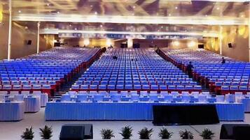 A large indoor auditorium with rows of blue seats, a stage at the front, and tables with white tablecloths.