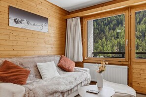 Une pièce chaleureuse avec un décor intérieur en bois, un canapé avec des coussins, une table blanche ornée d’un vase et une fenêtre donnant sur une forêt.