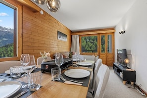 Un coin repas avec une table en bois dressée pour deux personnes, un téléviseur et une vue sur une zone boisée.