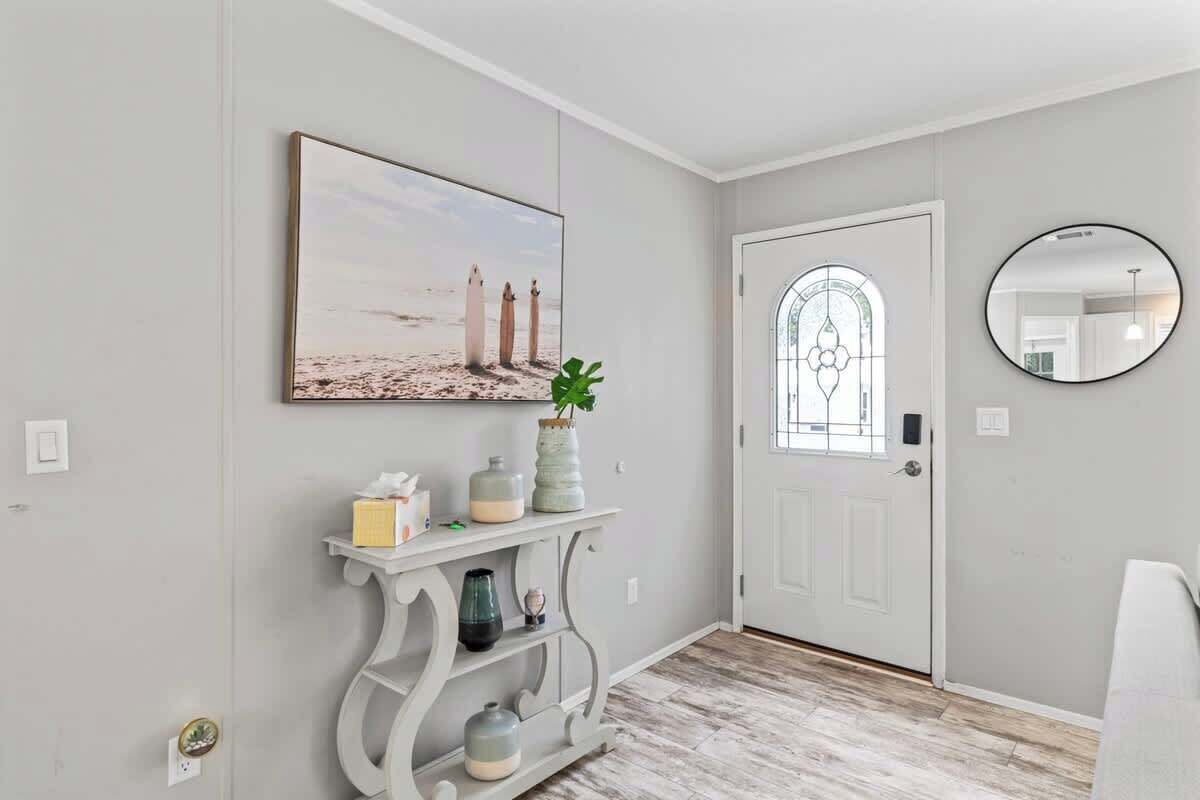 Un couloir avec une porte blanche, un miroir rond, une console ornée d’objets décoratifs et un tableau encadré accroché au mur.