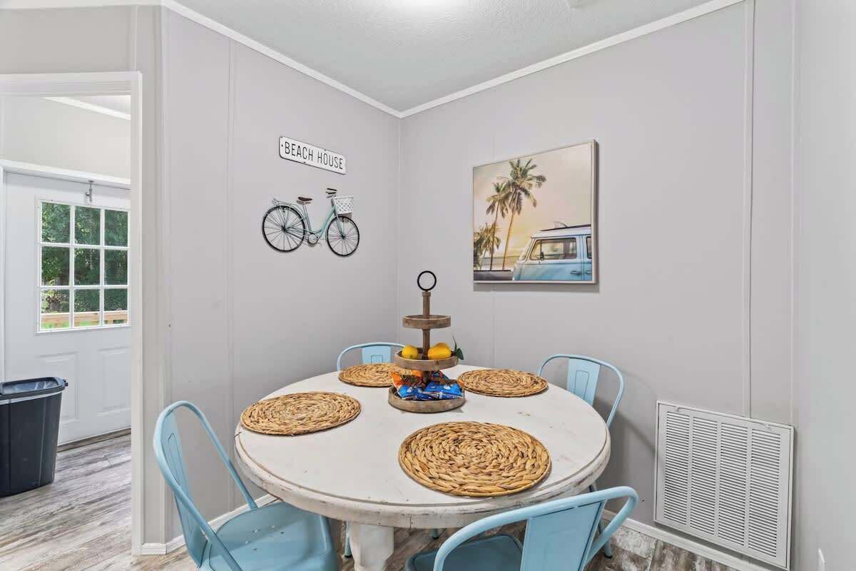 Een eetgedeelte met een ronde tafel, blauwe stoelen, geweven placemats, een fruitstuk als middelpunt, een fietsbordje aan de muur en een schilderij met een strandthema.