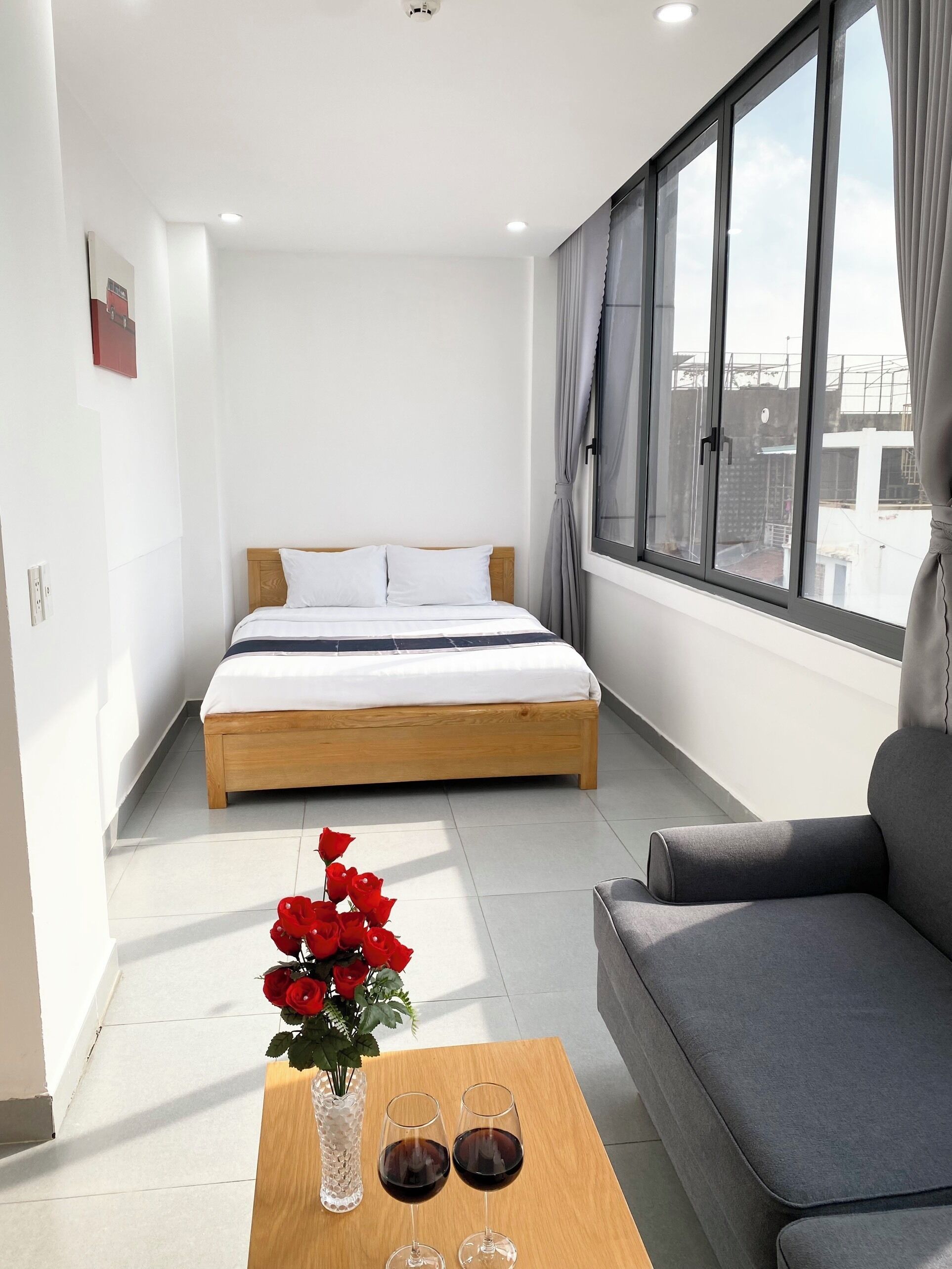 Une chambre d’hôtel moderne avec un lit, un canapé, une table ornée d’un vase de roses rouges et de deux verres de vin rouge, ainsi qu’une grande fenêtre avec des rideaux.