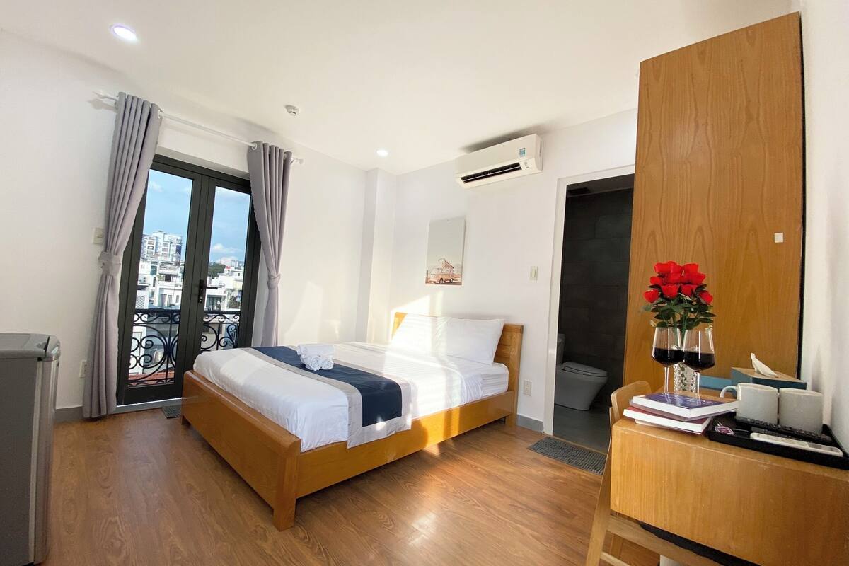 Habitación de hotel con cama de madera, un escritorio con un florero y vista a la ciudad desde un balcón.