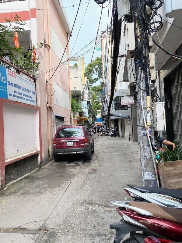 A narrow street with a parked red car and a motorcycle. There are buildings on both sides with visible signage and electrical wires overhead. - Home Away Minh Khai (Ho Chi Minh City)