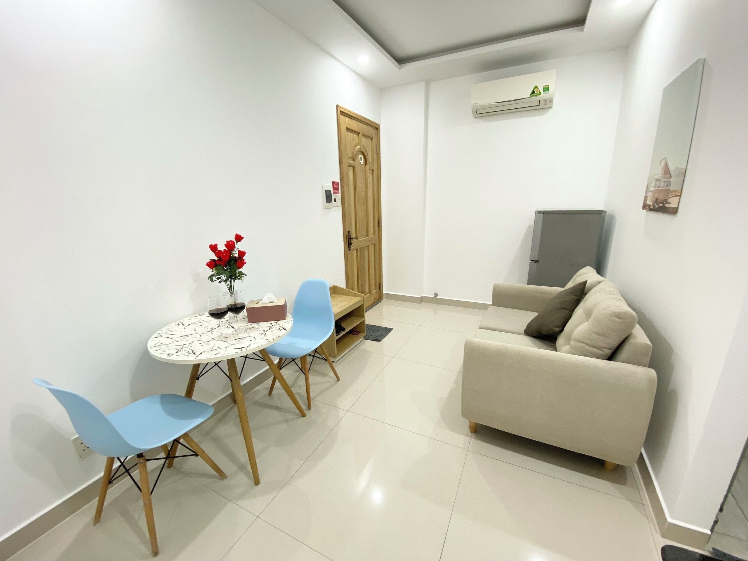 A small, modern living room with a beige sofa, a round table with two chairs, and a wooden door.
