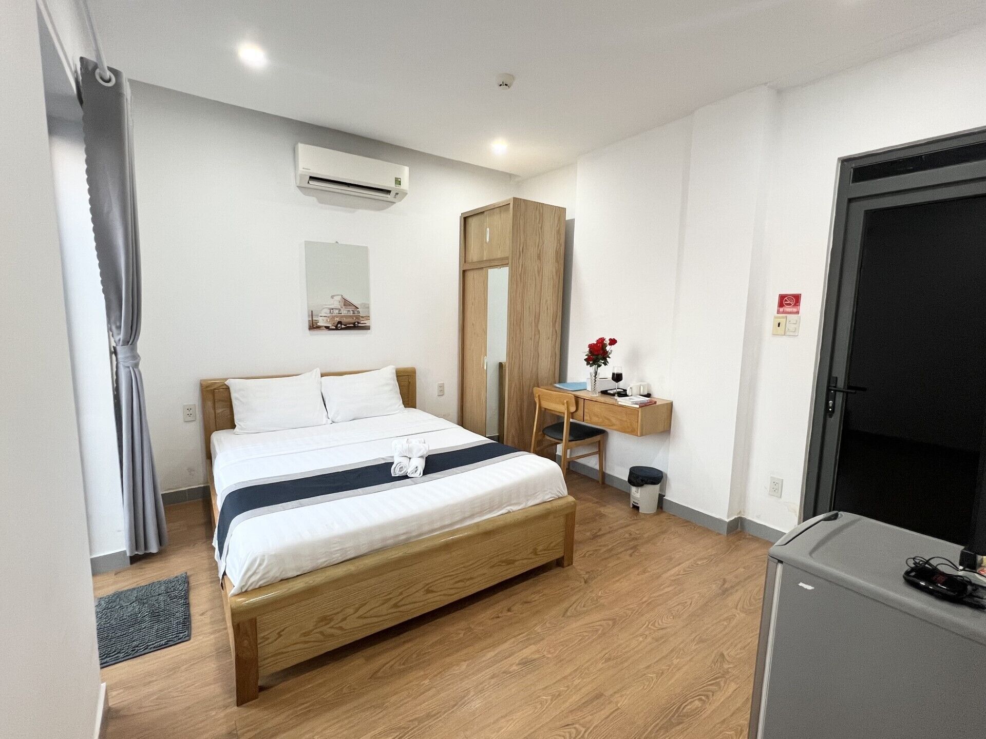 A hotel room with a wooden bed, a desk with a chair, an air conditioning unit, a wardrobe, and a door.