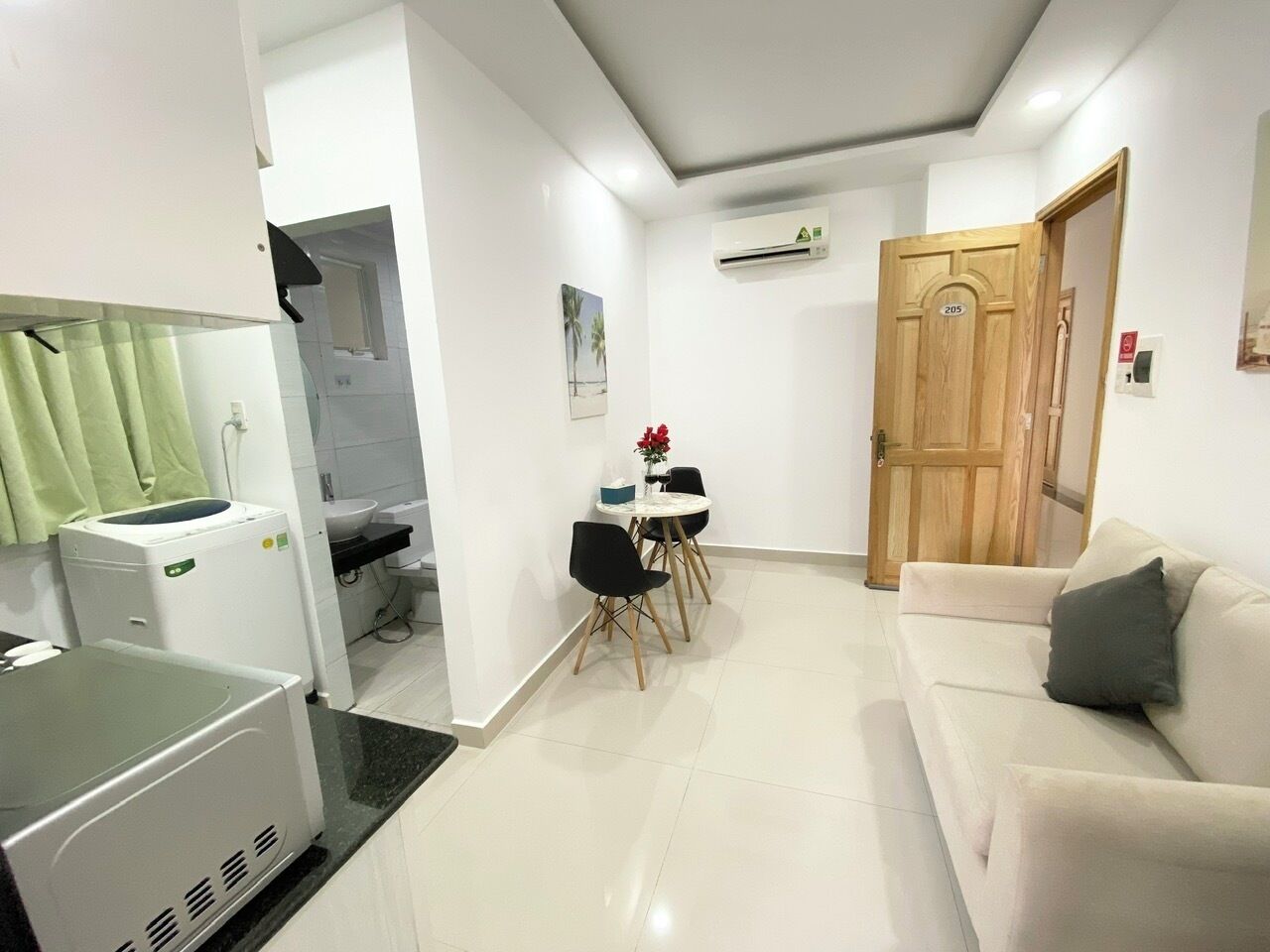 A modern living room with a white sofa, a dining table with chairs, a kitchen area with a washing machine, and a wooden door.
