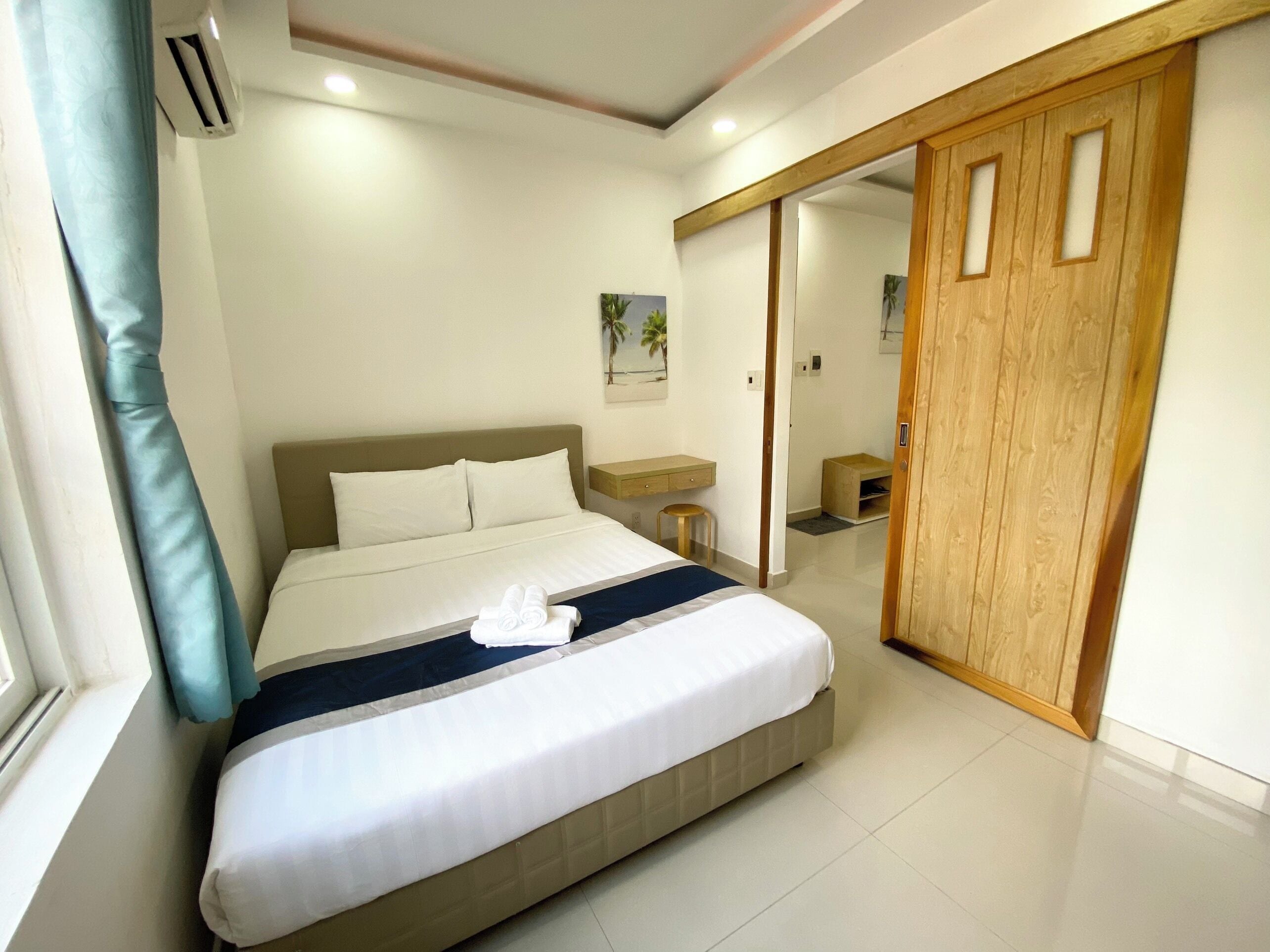 A neatly made bed with white linens and a blue accent, a wooden wardrobe, and a small wooden stool.