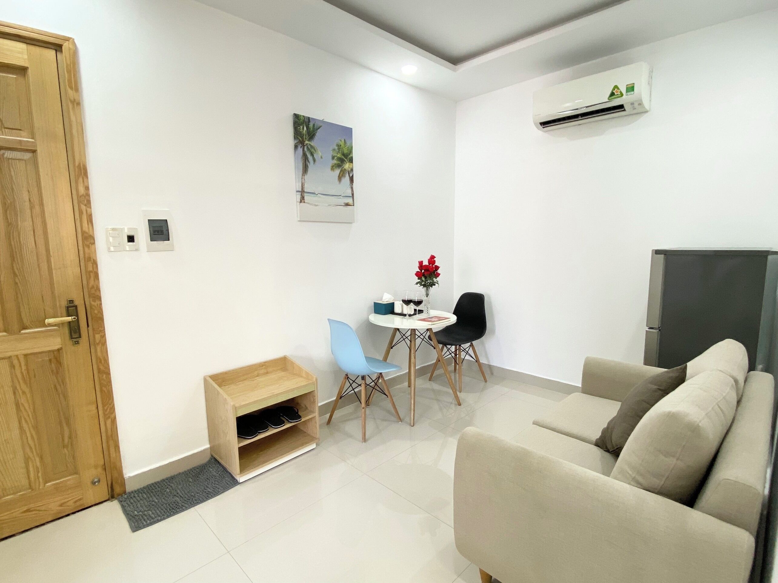 A modern living room with a beige sofa, a small dining table with two chairs, a wooden shoe rack, and a wall-mounted air conditioner.