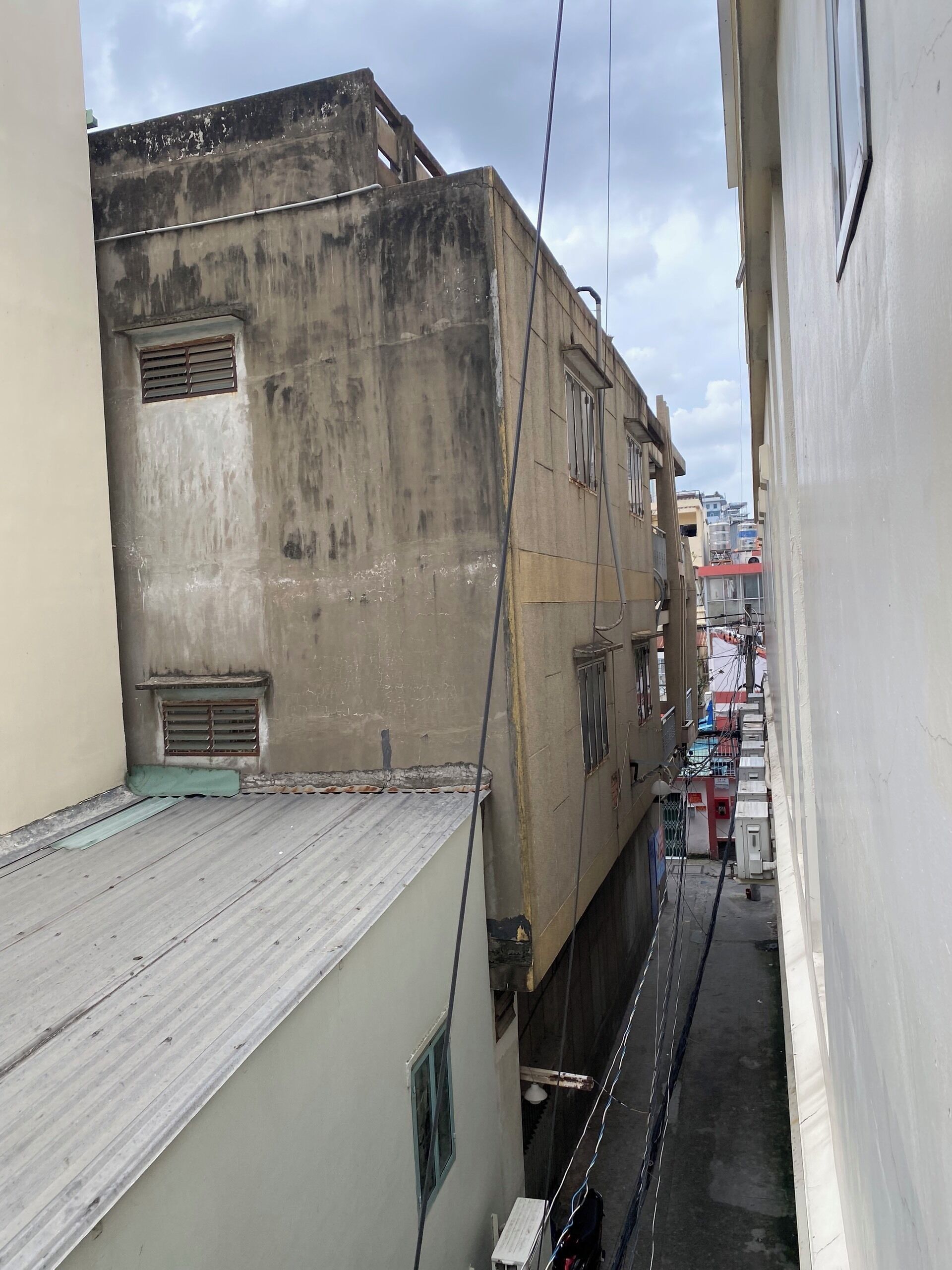 Un estrecho callejón entre edificios, con un muro de concreto en un lado y un edificio con techo metálico en el otro. Se observan cables y una pequeña rejilla de ventilación en el muro de concreto.