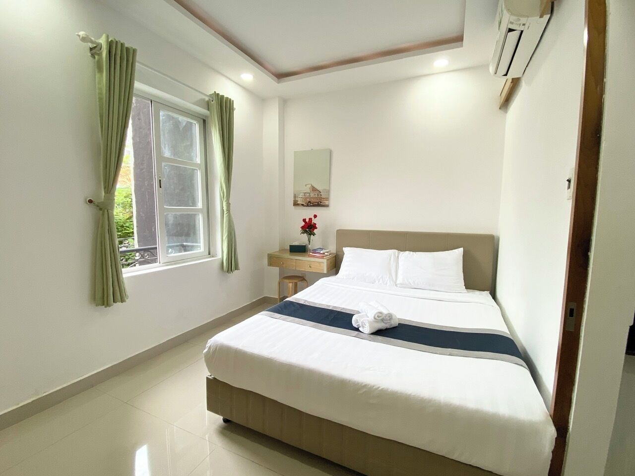 A neatly arranged hotel room with a bed, a bedside table, a window with curtains, and a wall-mounted air conditioning unit.