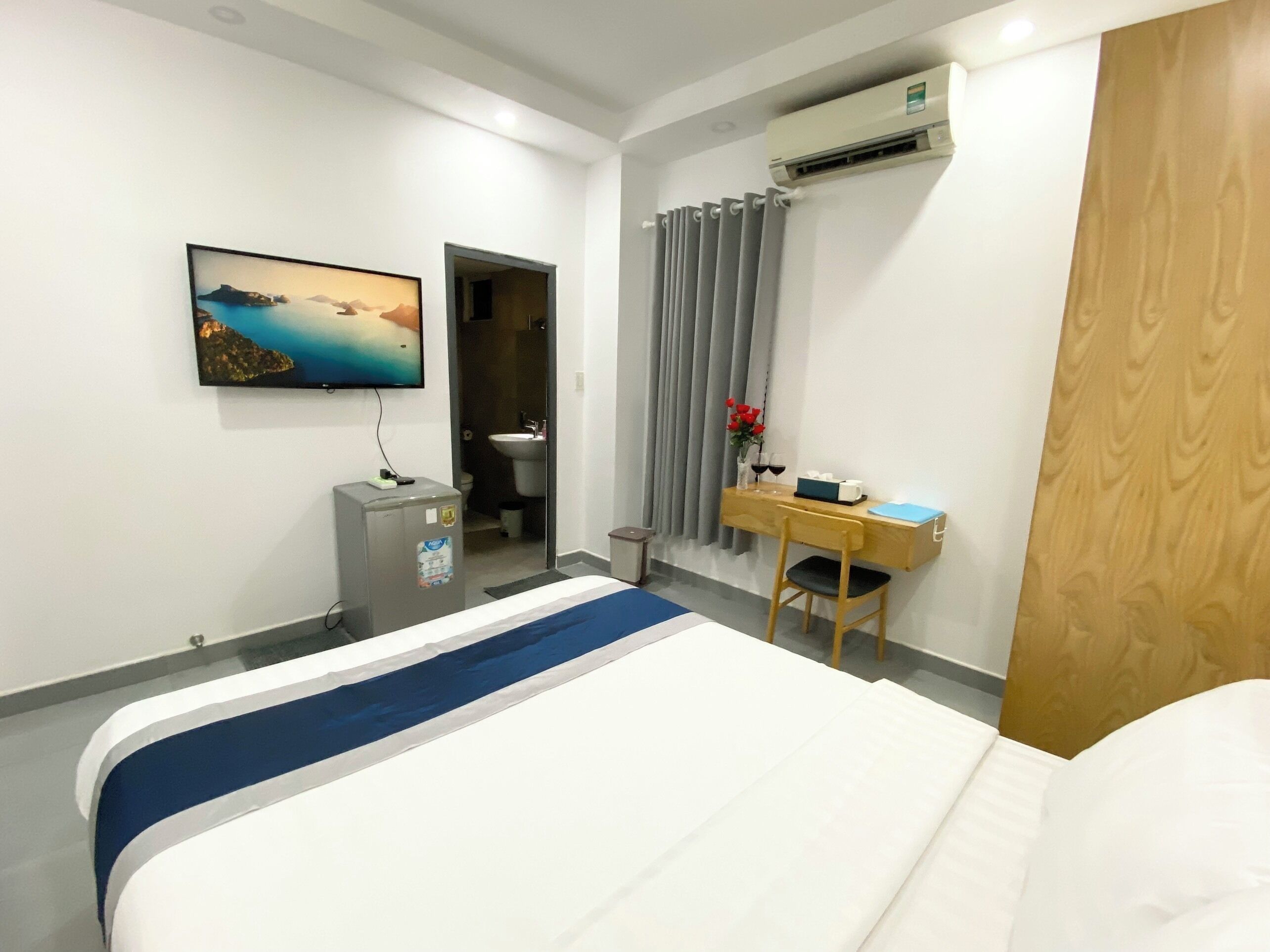 A hotel room with a bed, a desk with a chair, a small refrigerator, a bathroom visible through an open door, and a painting on the wall.