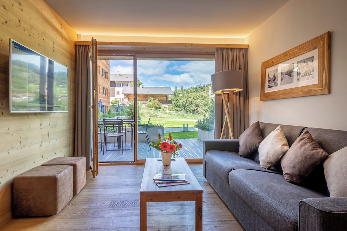 Una sala de estar con un sofá, una mesa de centro con un florero, y un ventanal con vistas a edificios y follaje.