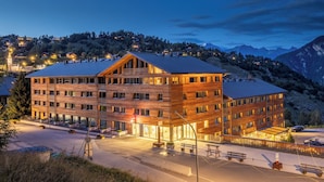 A multi-story hotel with a modern design, featuring a prominent entrance, outdoor seating, and a view of the surrounding mountainous landscape. - SWISSPEAK Resorts by Pierre & Vacances Premium Vercorin (Chalais)