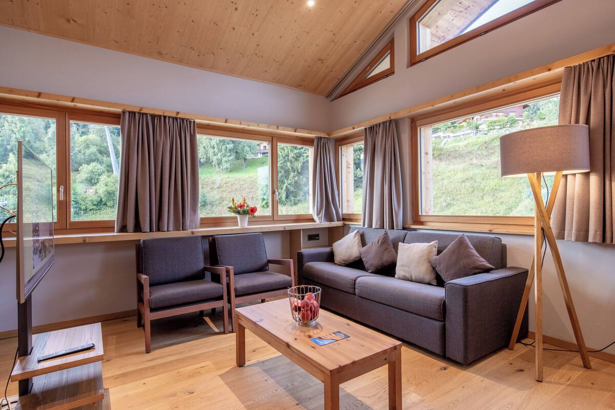 Una sala de estar moderna con sofá, sillas, una mesa de centro y ventanas amplias que ofrecen vistas a la vegetación.
