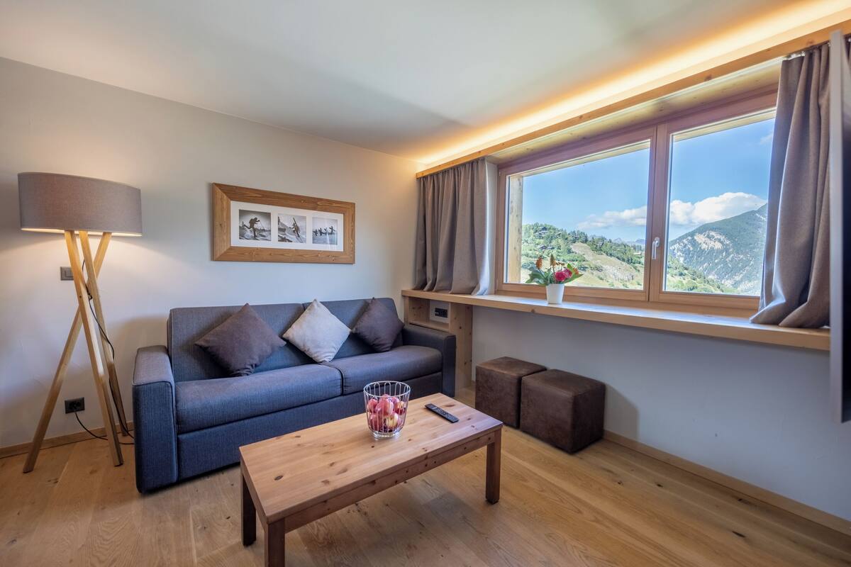 Una sala de estar moderna con un sofá, una mesa de centro con frutero, una lámpara y una ventana con vistas a montañas.
