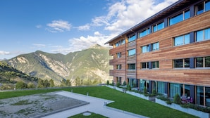 A modern building with large windows, a courtyard with grass, and mountains in the background. - SWISSPEAK Resorts by Pierre & Vacances Premium Vercorin (Chalais)