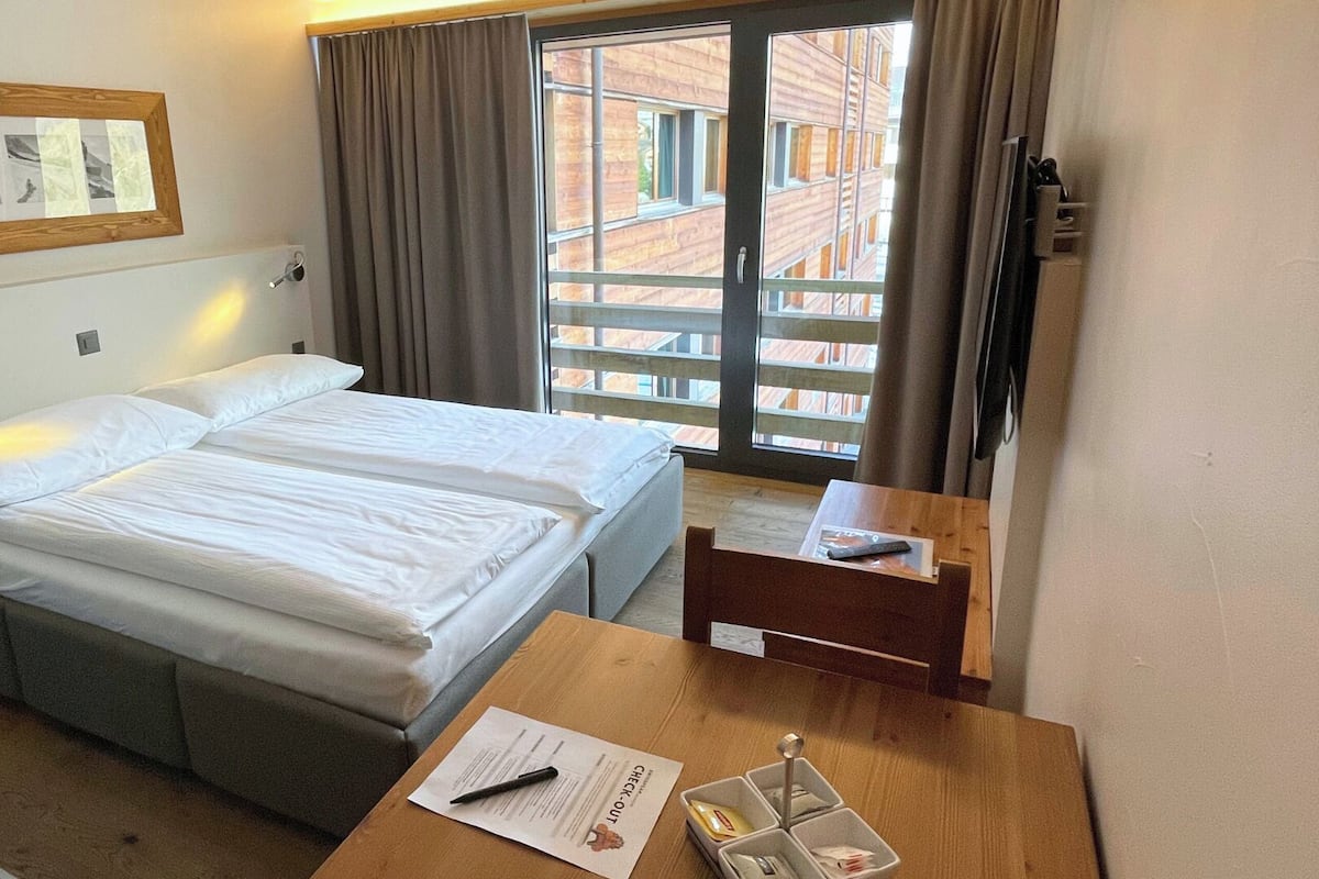 Una habitación de hotel con cama, escritorio con silla, ventana con cortinas, un cuadro en marco de madera en la pared y un cubo de basura pequeño.
