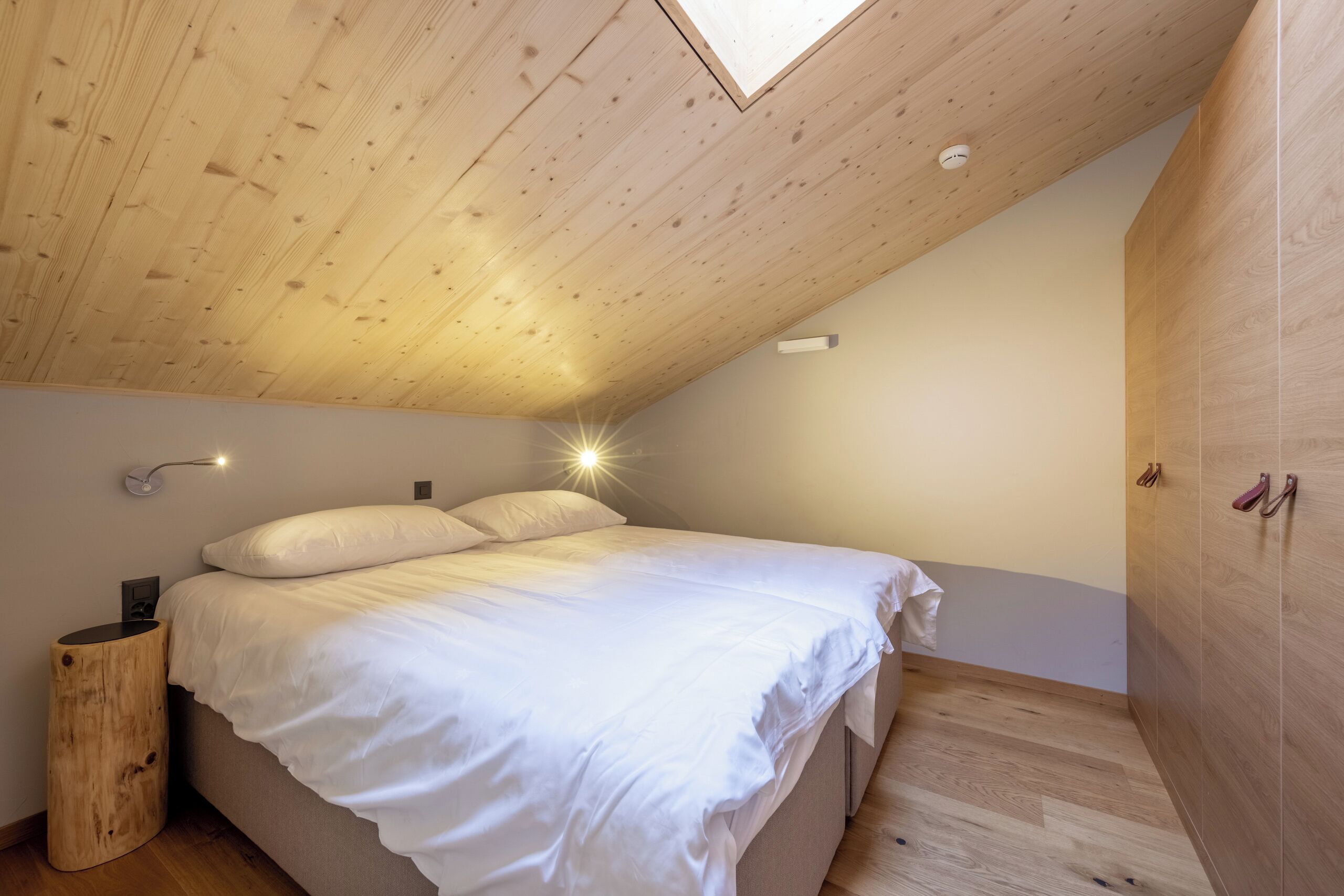 A bedroom with a sloped ceiling, a bed with white bedding, a bedside lamp, and a wardrobe.