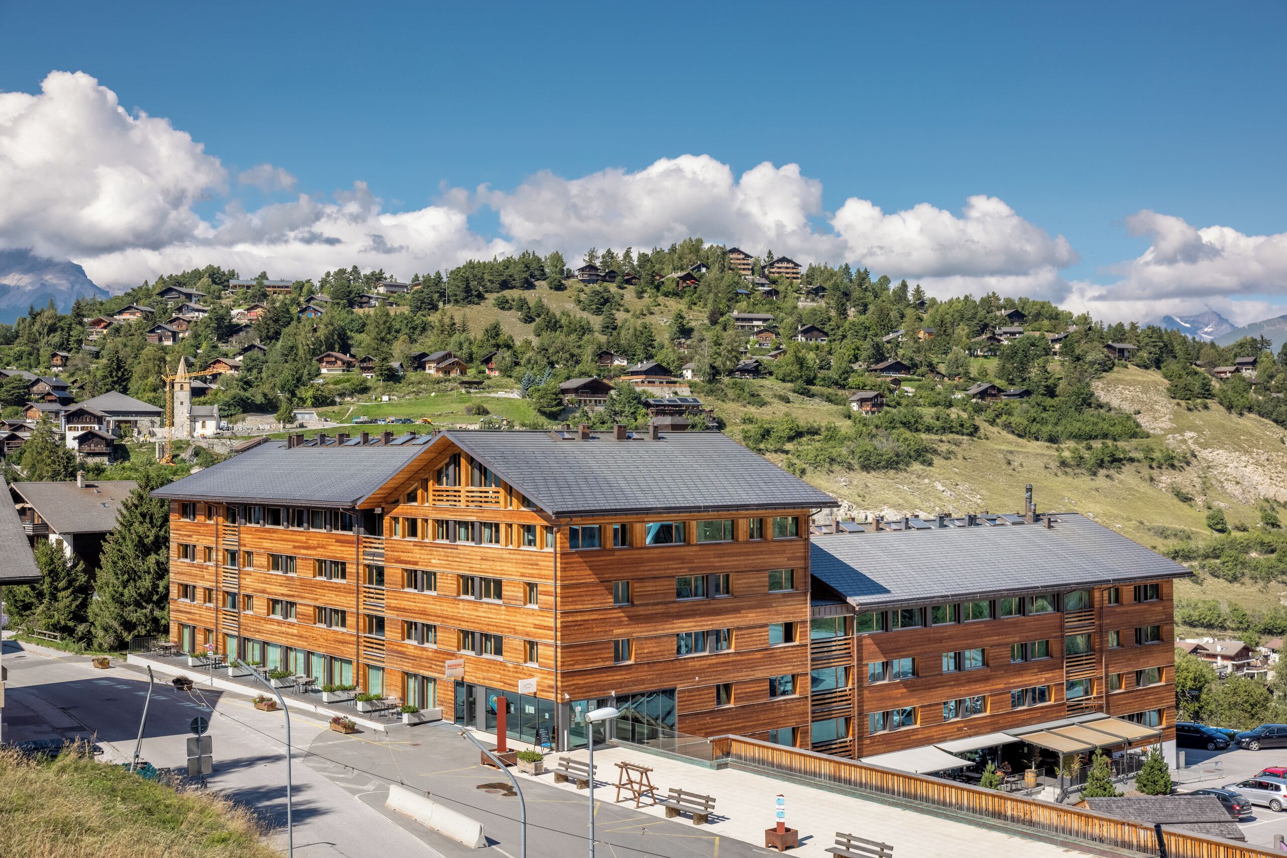 A multi-story wooden building with a flat roof, located in a mountainous area with visible houses in the background.