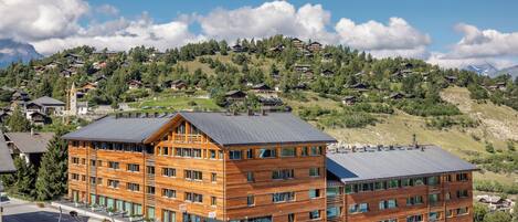 A multi-story wooden building with a flat roof, located in a mountainous area with visible houses in the background.