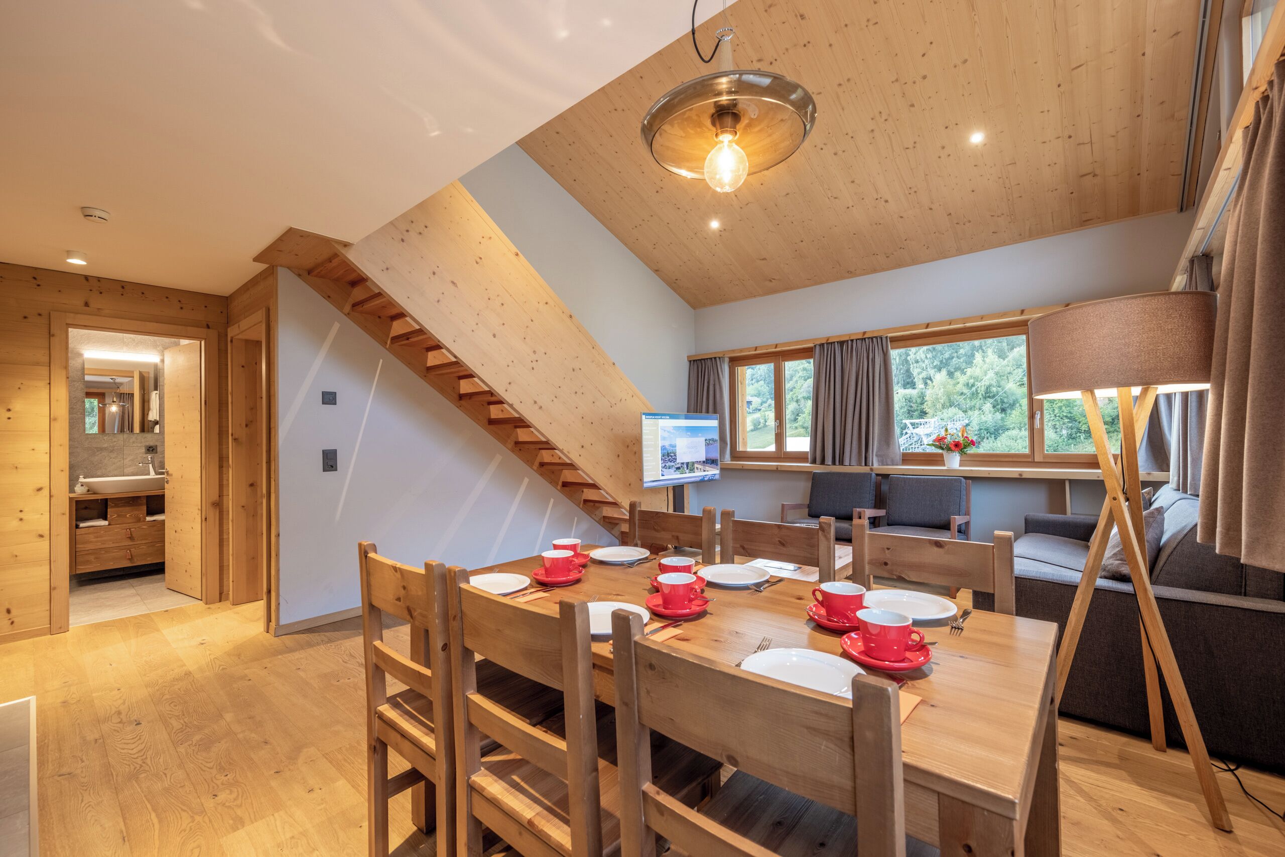 A wooden interior with a dining table set for four, a staircase, and a TV mounted on the wall.