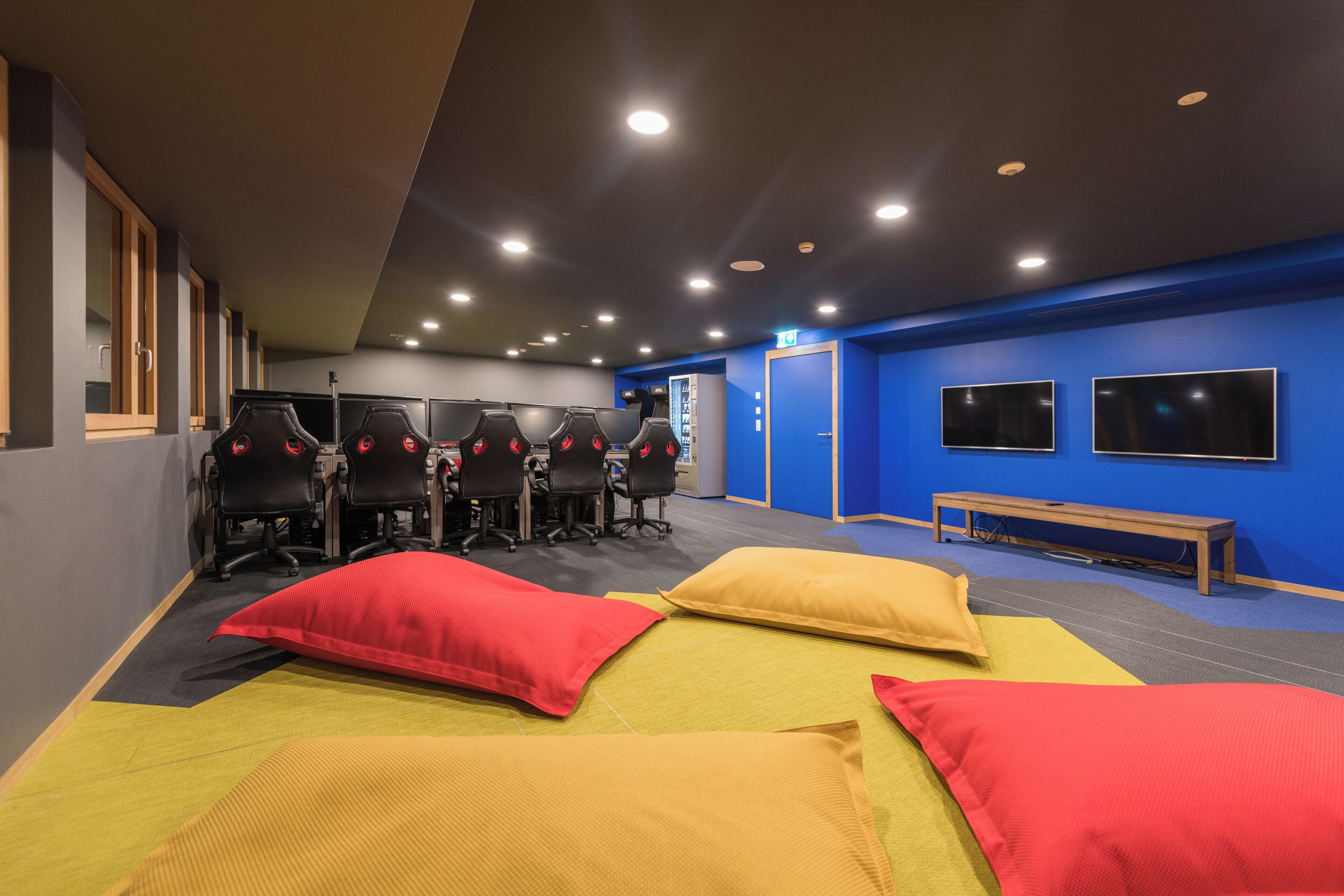 A room with four gaming chairs, a bench, and two large screens on the wall.
