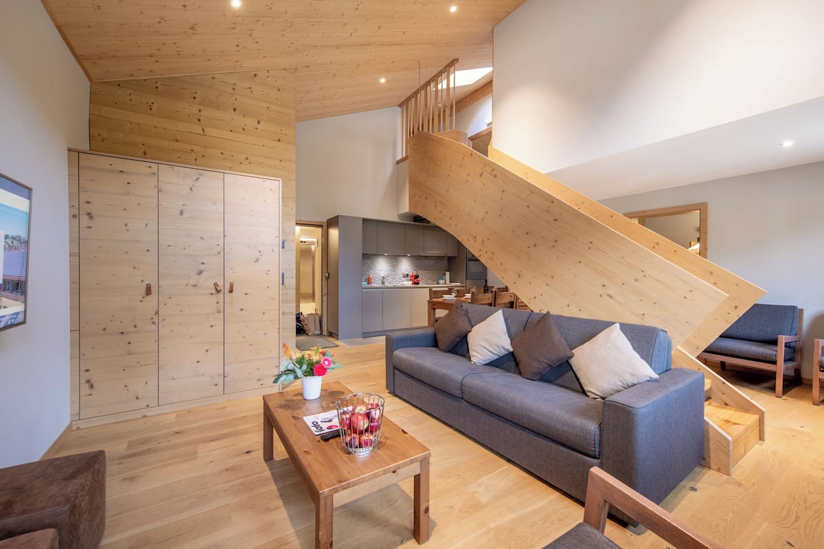 Una sala de estar moderna con una escalera de madera, un sofá gris, una mesa de centro de madera y una cocina al fondo.