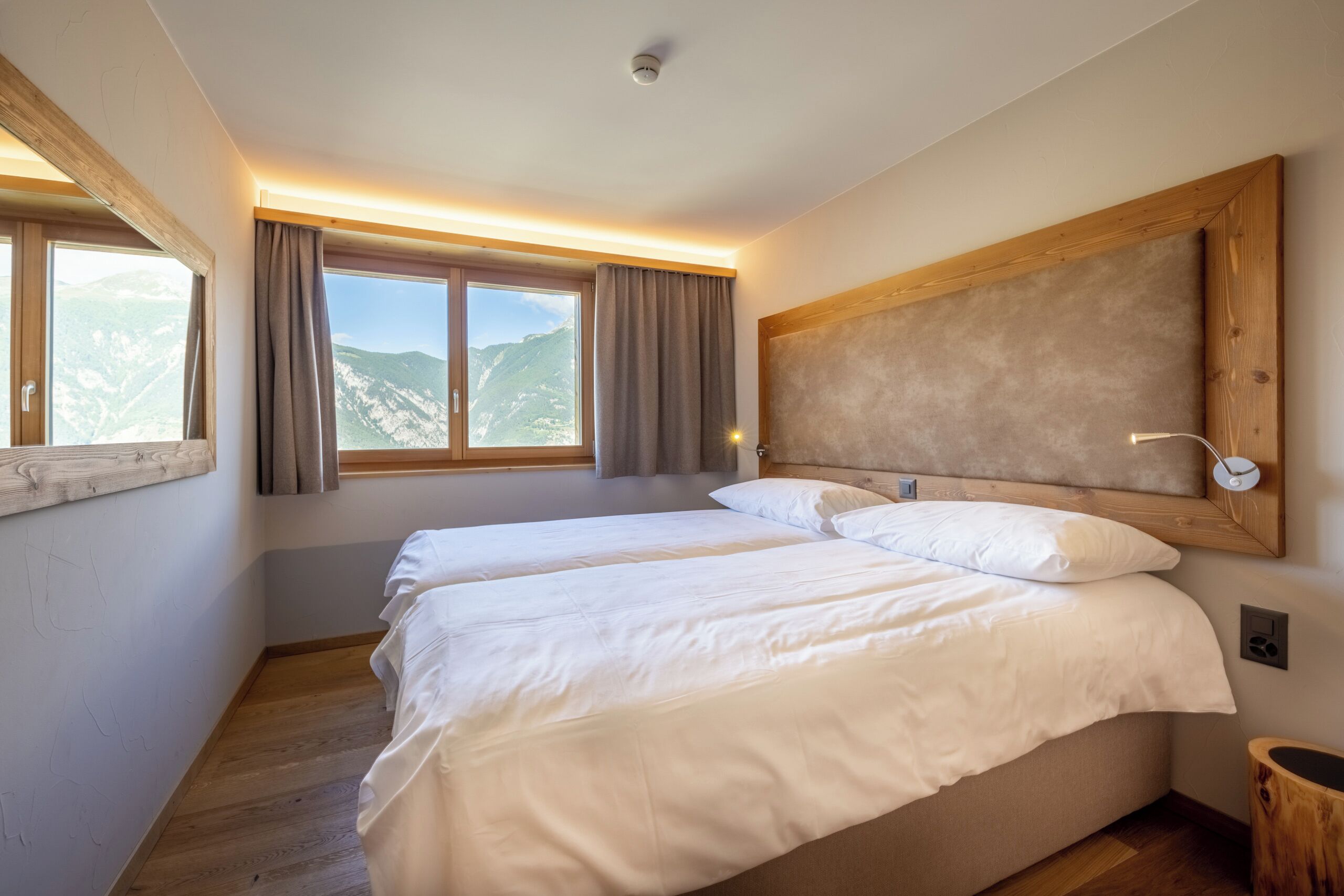 Un dormitorio con dos camas, un ventanal con cortinas, cabecero de madera y vistas a montañas.