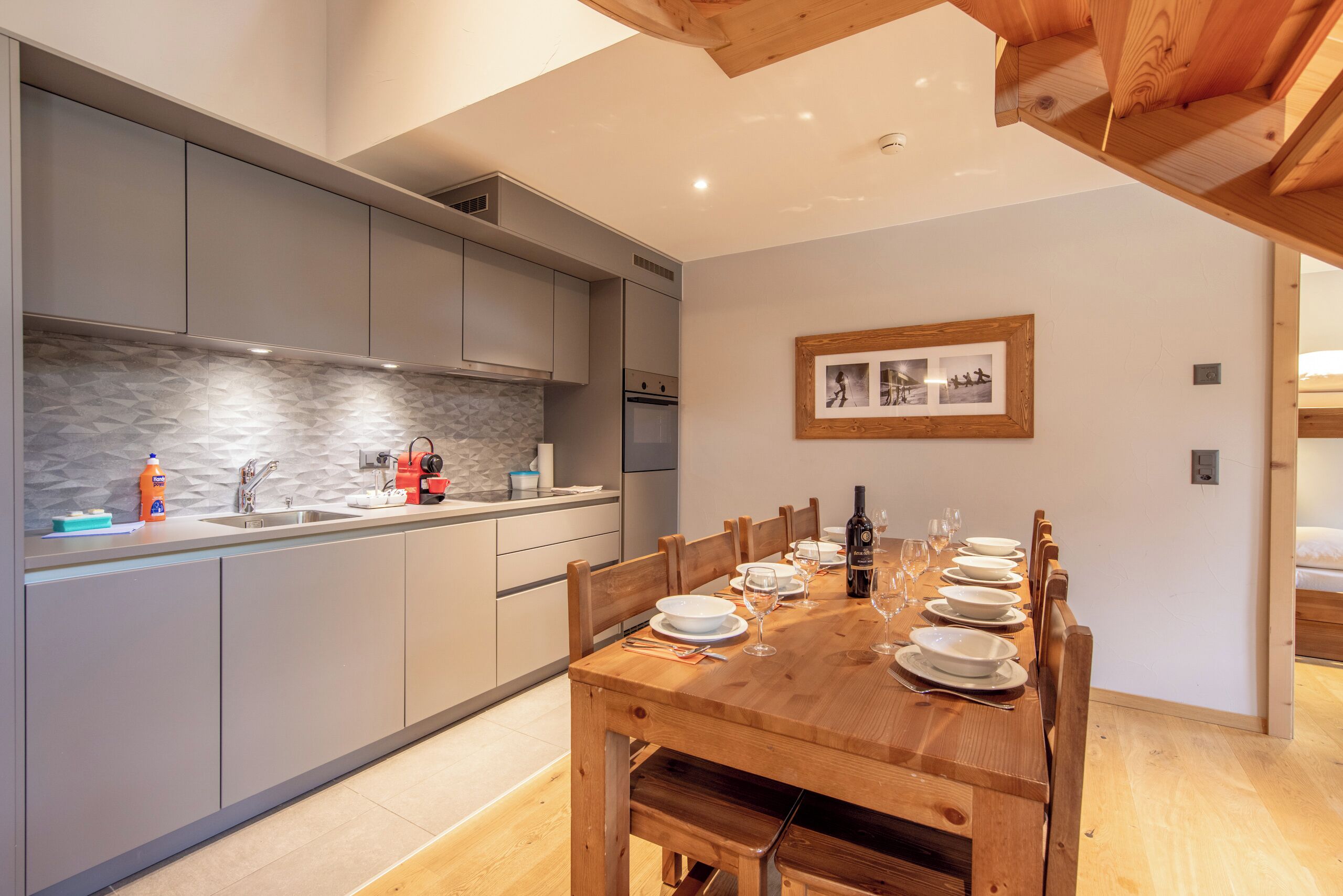 Una cocina moderna con armarios grises, una mesa de comedor de madera preparada para seis personas y un cuadro en la pared.